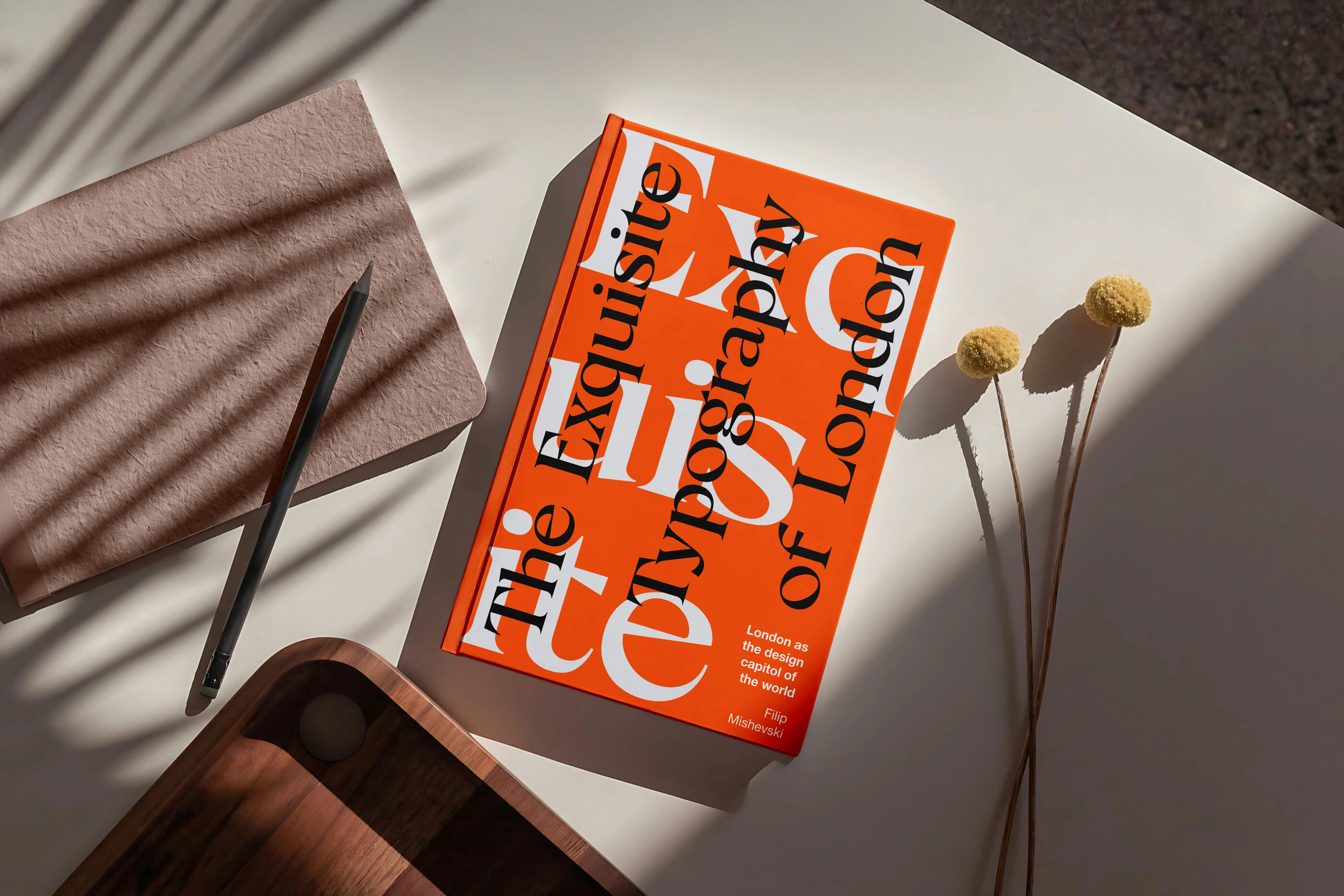 A mockup of the finished book on a sunlit table beside a pencil and dried
             flowers: an orange cover with The Exquisite Typography of London set in
             overlapping black and white serif letters.