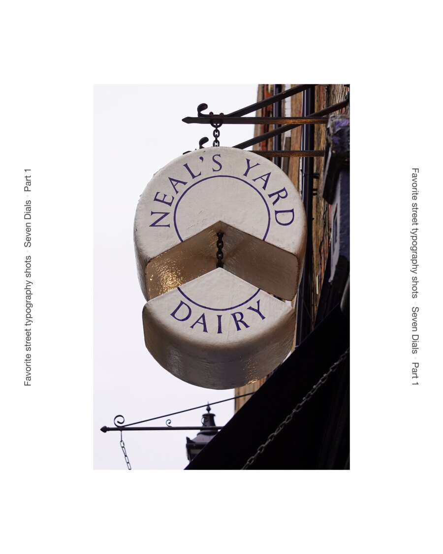 A hanging shop sign cut as a wheel of cheese with a wedge lifted out of it, lettered Neal&rsquo;s Yard Dairy in purple capitals around the rim.