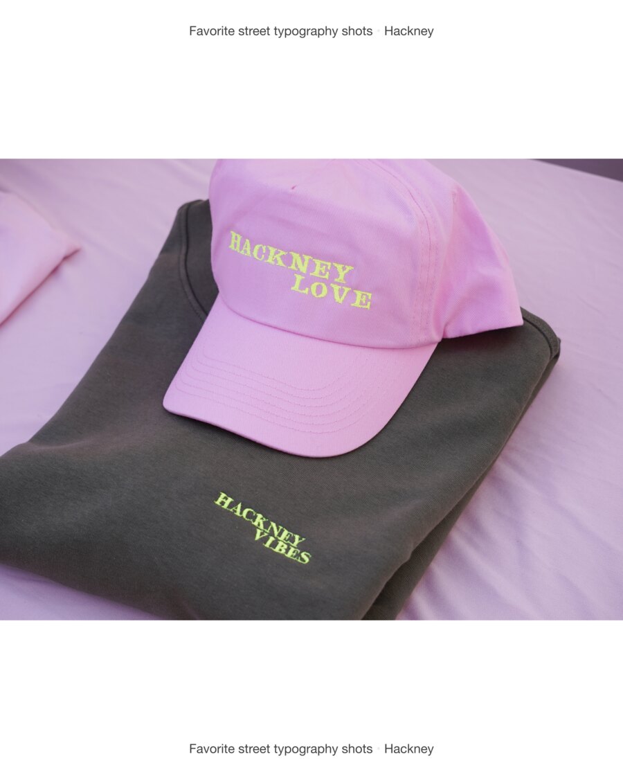 A pink cap embroidered Hackney Love in yellow-green condensed capitals, resting on a folded grey t-shirt embroidered Hackney Vibes in the same lettering.
