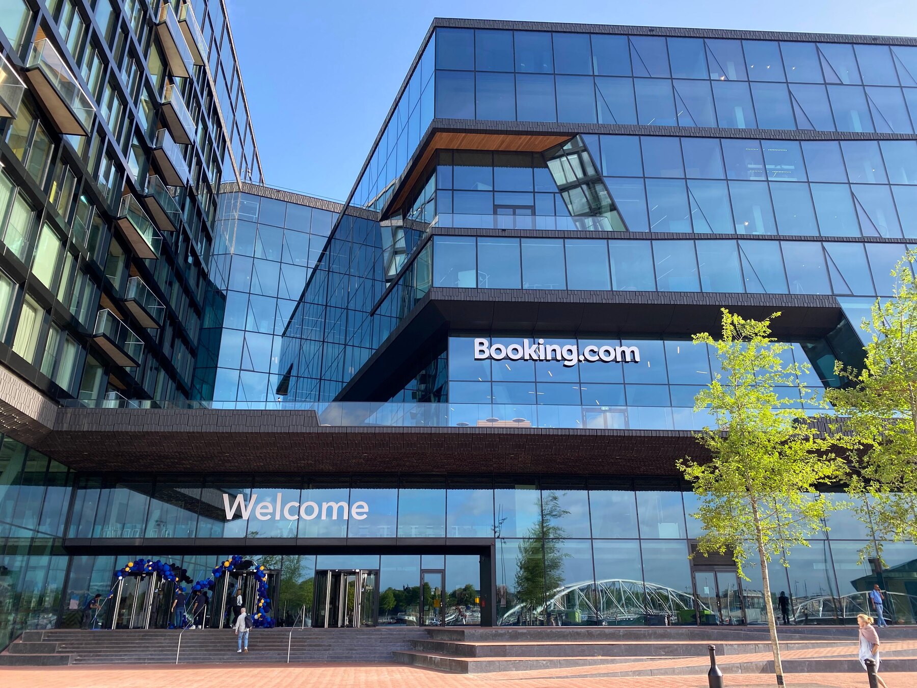 The glass front of the Booking.com headquarters in Amsterdam, the logo across the facade and the word Welcome on the window below it, with people walking in.